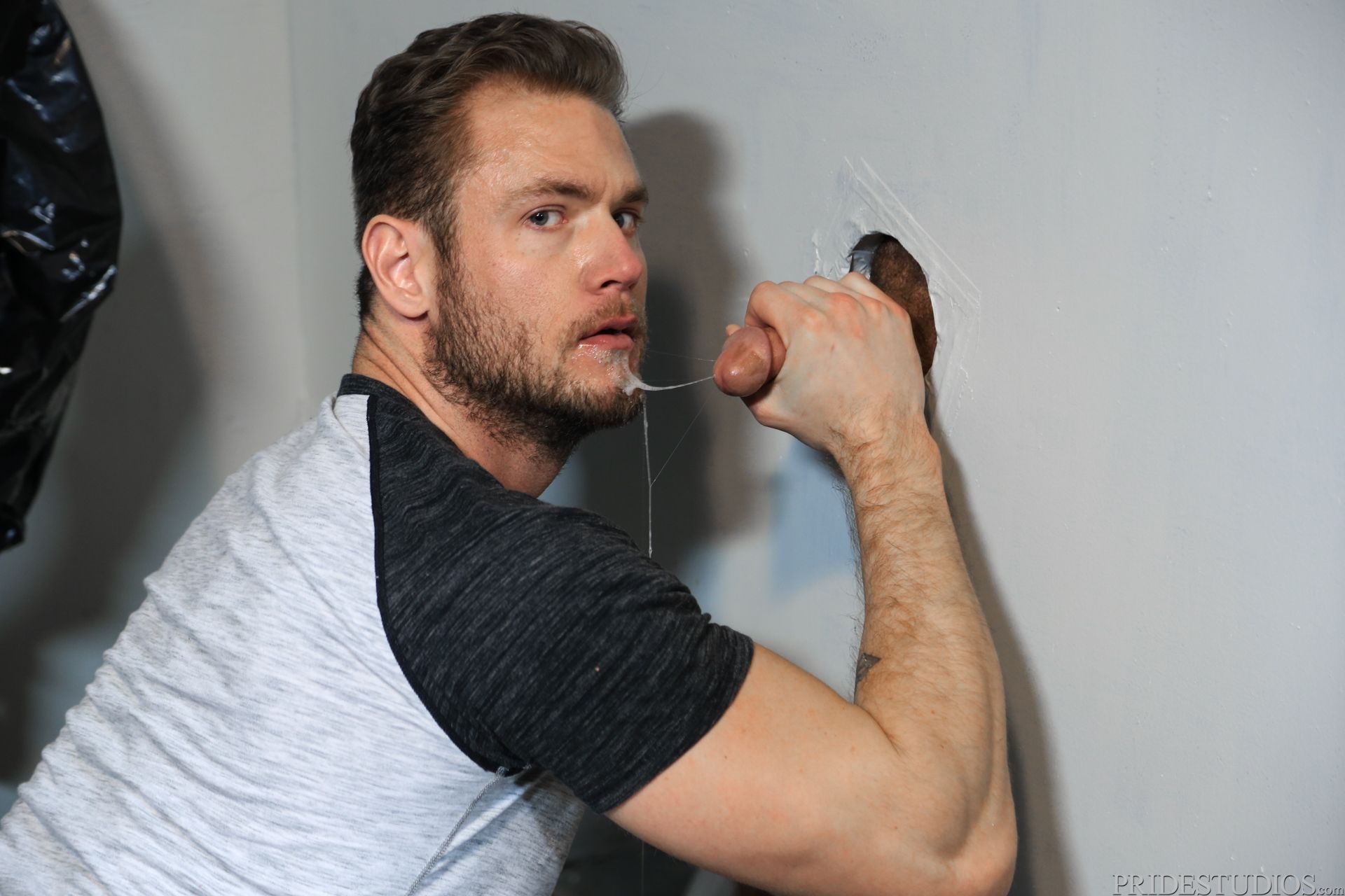 gay bearded male wearing t-shirt with his mouth covered in jizz and spit holding a thick cut cock at the local gay gloryhole